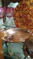 Glassware, hammered aluminum bowl, covered metal container, and decorative autumn leaf plate on glass shelf