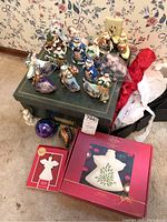 Photo displaying multiple hand-painted Thomas Kinkade Old World collection Santa Claus figurine ornaments arranged on a dark green box, with two Lenox boxed ornaments and some loose packing material around.