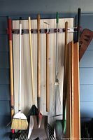 Full view of gardening tools including rakes, shovels, broom, and hoe hung on storage hooks against wall.