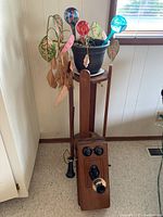 Photo showing wooden Kellogg hand crank phone with plant stand holding live plant and glass watering bulbs.