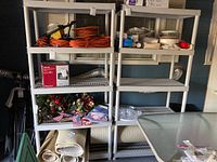 Wide view of a gray plastic shelving unit stocked with various household items including orange extension cords, Christmas decorations, rolled area rugs, and kitchen supplies. Unit shows scuffs and scratches.