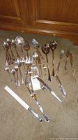 Silverware assortment including spoons, forks, knives, serving utensils laid out on carpet in front of wooden cabinet.