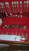Top rack of wooden chest displaying 12 dinner knives with ornate handles seated in red velvet slots.