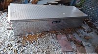 Full front and top view of large aluminum diamond plate tool chest labeled Truck Trunk, resting on gravel ground with some debris around.