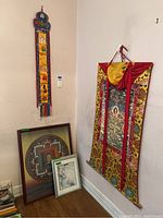 Wide view showing a long narrow textile wall hanging with Tibetan script on the wall, large textile wall hanging with detailed traditional art, and framed circular mandala print leaning against the wall, plus smaller framed print.