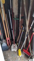 Photo showing shovel, post hole digger, weed puller, brooms, mop, and garden rake grouped together in a corner.