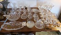 Overview of several clear glass and lead crystal goblets, glasses, serving bowls, a glass cloche, and candlestick holders arranged on a wood table showing variety of styles, shapes, and etched detailing.