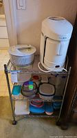 Metal rolling cart with three shelves containing a white Baby Blend appliance, white salad spinner with handle, assorted plastic and glass food storage containers in various colors and sizes.