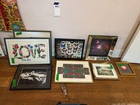 View of all eight framed pieces arranged side by side against a wall, showing a mix of needlepoint and prints including butterfly, floral, photograph, and abstract art