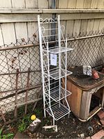 Front view of white metal 4-tier plant stand showing tiers and decorative scrollwork, standing outside by a chain-link fence.