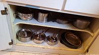Cabinet interior showing various pots, pans, and lids including stainless steel, aluminum, and glass lids.