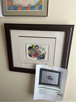 Framed multicolored mono print painting titled 'Garden' by Lloyd Walter Bennett Junior, shown hanging on a wall with a partial view of another colorful artwork above.