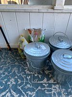 Photo of three aluminum trash cans with lids and several birdseed bags on a carpeted floor near a white wall.