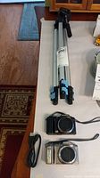 Folded metal tripod with blue locks lying flat on a table next to the two Olympus digital cameras, one silver and one black with wrist straps and a connected cord.
