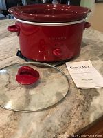 Red oval Crock Pot slow cooker displayed on countertop with unplugged power cord, rotary dial, glass lid with red handle, and instruction manual.