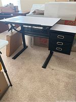 White drafting table with black metal frame, angled legs, and mesh shelf beneath. The tabletop is rectangular and white with a broken lift mechanism. On the right side, there is a three-drawer black metal cabinet with silver handles.