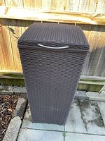 Tall brown plastic wicker style wastebasket with attached lid shown outdoors near a wooden fence.