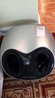 Top view of silver and black foot massager showing openings for feet and attached power cord inside one opening