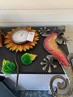 Photo of metal lizard wall art and sunflower-shaped thermometer together on a black surface.