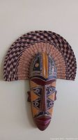 Front view of the West African mask showing painted geometric patterns in various colors and raffia fan accent.
