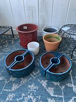 Photo showing six planters of various colors and materials, including blue ceramic semi-circle pots, a large red ceramic pot, and a metal plant stand.