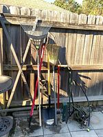Photo showing assortment of yard tools including rakes, shovels, broom, hand cultivator against wooden fence