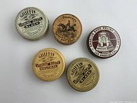 Five antique advertising tins on flat white surface showing replacement labels and metal lids, including shoe polish and leather soap tins in various colors of cream, tan, brown, maroon and white.