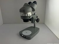 Angled full view of the microscope showing the binocular eyepieces, vertical focus and height adjustment stand, and base with white circular specimen disk.
