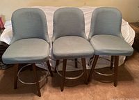 Front view of the three upholstered bar stools arranged side by side showing the light blue/grey fabric seats and backs with silver nailhead trim.