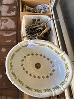 Top-front view of oval ceramic sink with floral hand-painted decoration and boxes containing brass plumbing accessories behind.