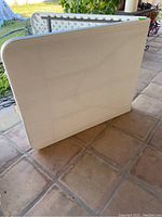 Full view of the white folding table closed and standing on end, top surface visible with rounded edges.