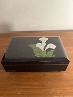 Closed view of rectangular black lacquer box with painted white calla lilies on top, placed on wood surface against light wall.