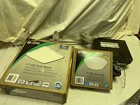 Photo shows two boxed light fixtures labeled Commercial Electric with product labels for 11" LED Flush Mount and 7" LED Versi Lite, plus part of a metal light fixture with frosted glass shades behind the boxes.