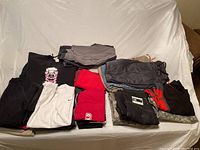 Photo of folded men's clothing including sweatshirts, pants, shorts, and shirts spread on white cloth.