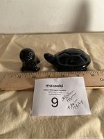 Two small black crystal figurines placed on beige cloth with ruler showing size. One is a turtle and the other is a baby seal. Both pieces are highly polished and smooth.