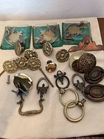 Clear view of various vintage brass cabinet pulls, knobs, and handles spread out on a flat surface including those in packaging and loose pieces.