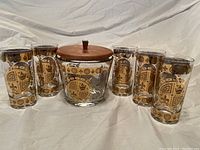 Front view of the ice bucket with wooden lid surrounded by six matching glasses with gold decorative pattern.