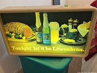 Frontal view of the illuminated Lowenbrau bar sign showing the full graphic with bottles, beer mug, food items and slogan.