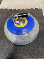 Photo showing a single used granite curling stone with a blue and black handle and yellow ring around the handle base. The curling stone has visible wear and surface marks from use.