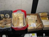 Photo showing five trays and boxes filled with new vintage clock parts, mostly sealed in envelopes and plastic bags organized for sale or inventory.