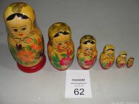 Six vintage Russian Matryoshka dolls arranged by size from largest to smallest on a white surface with auction lot card number 62 in front.