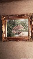 Framed artwork showing a house with a thatched roof by a stream, with a small stone bridge in front, and trees in background.