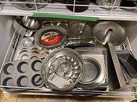Wide view of various metal baking tins, pans, trays including muffin tray, pudding tins, loaf pan, and pie tins piled in a shelf