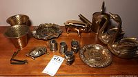 Overview of all brass objects on wooden surface including bowls, ashtray, piggy bank, swans, watering can, turtle container, small cups, and small stand.