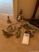 Overview of all brass items showing the hand bell, bird figurine, horseshoe door knocker with seated figure, bell with chain door knocker, and two candlestick holders.