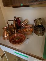 Copper kitchenware including tea pot, baking molds, colander, and decorative baskets arranged on a counter