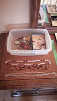 Plastic container holding various brooches, pins, and loose pearls, placed on a wooden surface with three necklaces laid out in front.