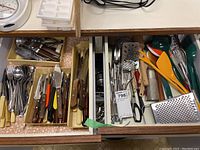 Drawer with assorted knives, forks, spoons, graters, scissors, peelers, orange plastic spatulas, and masher.