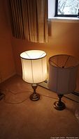 Two table lamps placed on carpet near window with curtains, showing lamp bases and shades, one lamp illuminated showing internal structure and fabric damage.