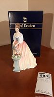 Frontal view of Royal Doulton Alexandria figurine with woman watering flowers and original box in background.